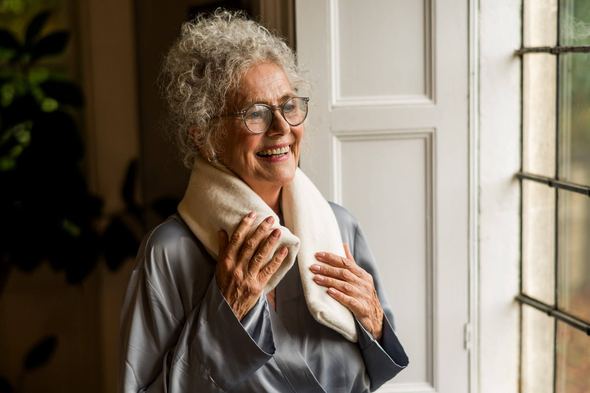 A woman wearing a cream CosyPanda long hot water bottle around her shoulders beside a window